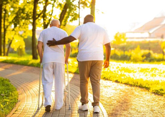 Caregiver is teaching old man to walk with walker. Professional nurse and patient walking outdoor at sunset. Assistance, rehabilitation and health care.